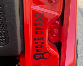 May include: Close-up of a bright red vehicle door frame. Black text reads "NAKED MODE" vertically, with an "ON" indicator below. The door frame features hinges and bolts.