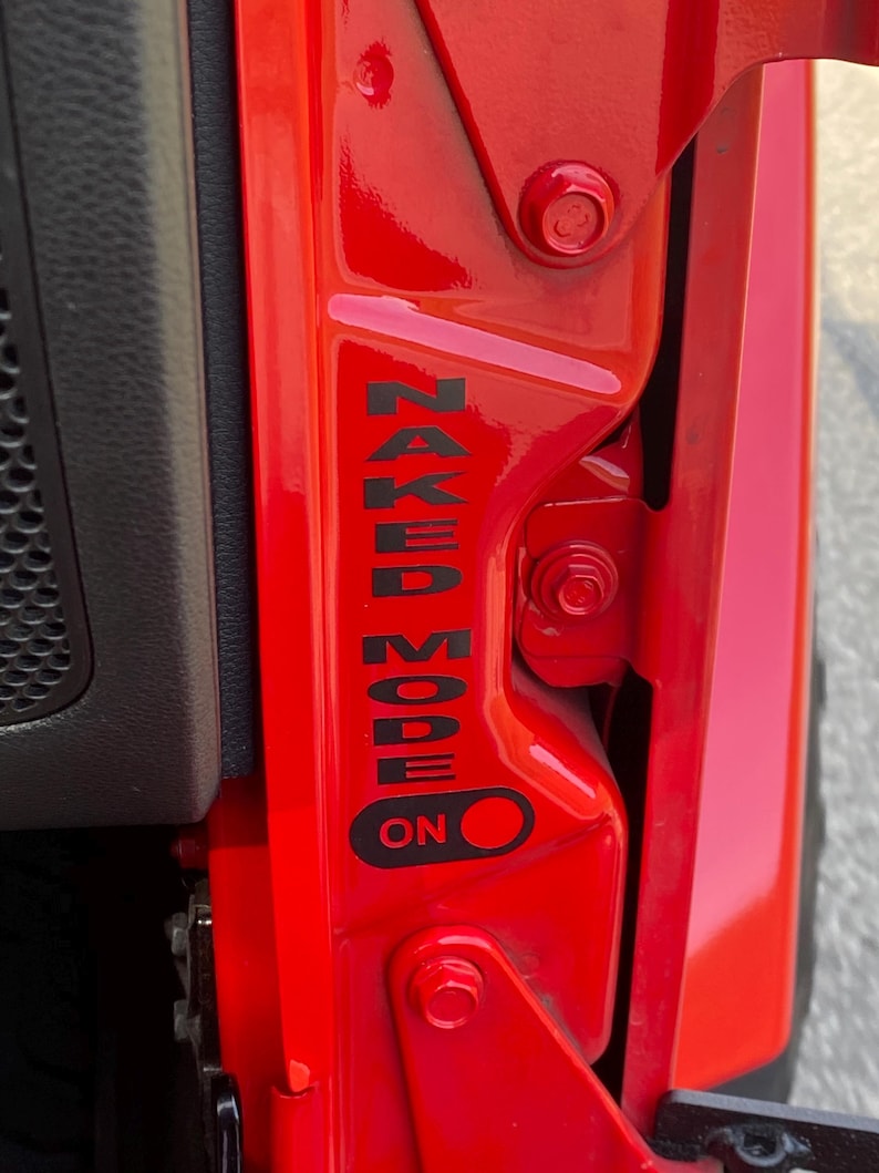 May include: Close-up of a bright red vehicle door frame. Black text reads "NAKED MODE" vertically, with an "ON" indicator below. The door frame features hinges and bolts.