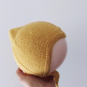 May include: A yellow knitted baby bonnet with a long tie. The bonnet is made of a soft, textured yarn and has a simple, classic design.