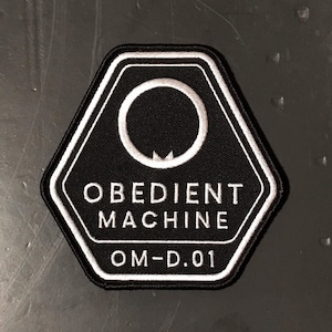 May include: Black and white hexagonal patch with the text "Obedient Machine" and "OM-D.01" in white. A white circle with a small white shape at the bottom is in the center of the patch.