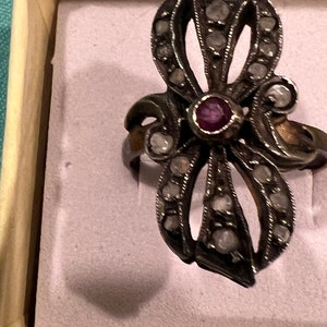 May include: A silver ring with a ruby gemstone in the center and a bow design with small diamonds around it.