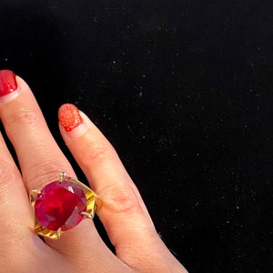 May include: A gold ring with a large, round, red gemstone. The ring is on a person's finger.