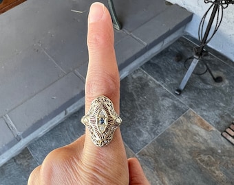 May include: A gold filigree ring with a blue and white stone setting. The ring is on a finger.