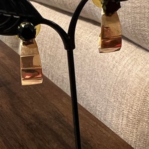May include: A pair of gold hoop earrings with black gemstone accents. The earrings are displayed on a black metal stand with leaf-shaped arms.