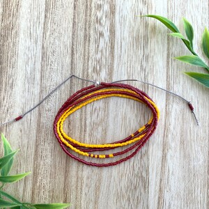 May include: A multi-strand beaded necklace with alternating red and yellow beads. The necklace is tied with a gray cord.