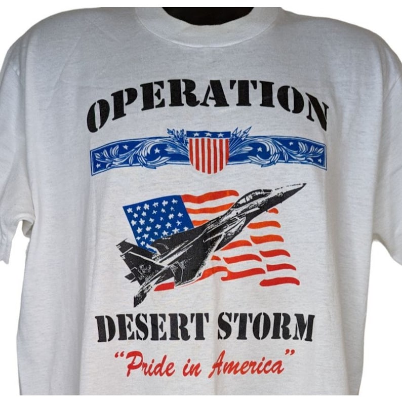 May include: White t-shirt with a black and white image of a fighter jet flying over an American flag. The text "OPERATION DESERT STORM" and "Pride in America" is printed on the shirt.