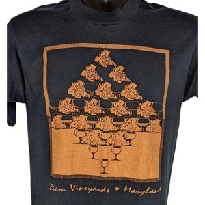 Può includere: Maglietta nera con una grafica marrone di una piramide di uva e bicchieri di vino. La grafica è circondata da un bordo quadrato marrone. Il testo "Ziem Vineyards - Maryland" è stampato sotto la grafica.