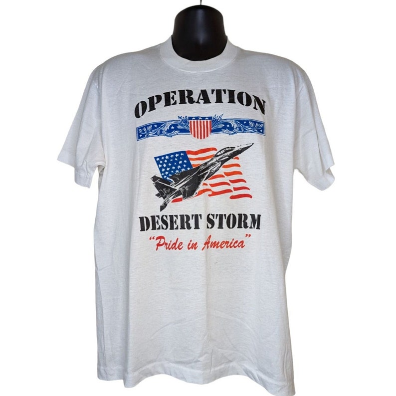 May include: White t-shirt with a black print of an F-16 fighter jet flying over an American flag. The text "Operation Desert Storm" and "Pride in America" are printed below the image.