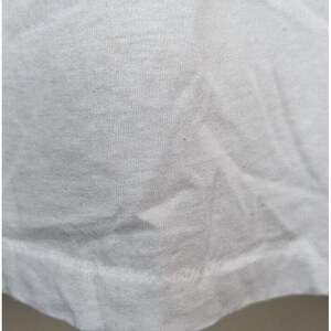 May include: Close-up of a white cotton t-shirt fabric. The fabric has a smooth texture and a slightly wrinkled appearance.