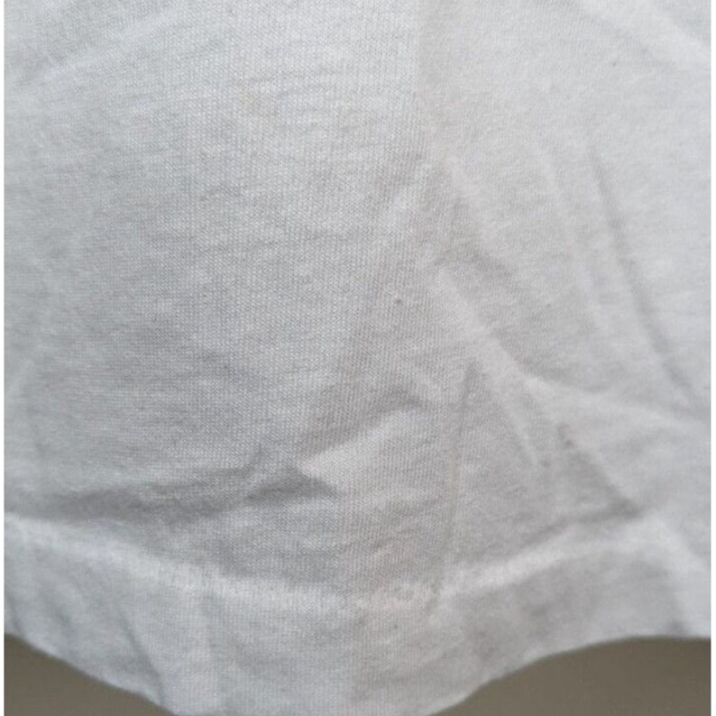 May include: Close-up of a white cotton t-shirt fabric. The fabric has a smooth texture and a slightly wrinkled appearance.