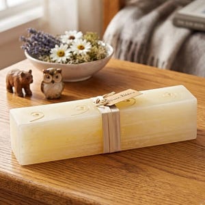 May include: A rectangular, cream-colored candle with decorative swirls and a tag that reads "Vance Kitira." The candle is wrapped with a beige ribbon. A small wooden bear and owl figurine sit next to a bowl of flowers.
