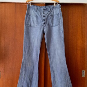 Puede incluir: Pantalones acampanados de mezclilla azul claro con bragueta abotonada y dos bolsillos delanteros. Los pantalones cuelgan de una percha de madera. La tela tiene un aspecto descolorido, lo que le da un aspecto vintage. Un estilo de pantal&oacute;n cl&aacute;sico.