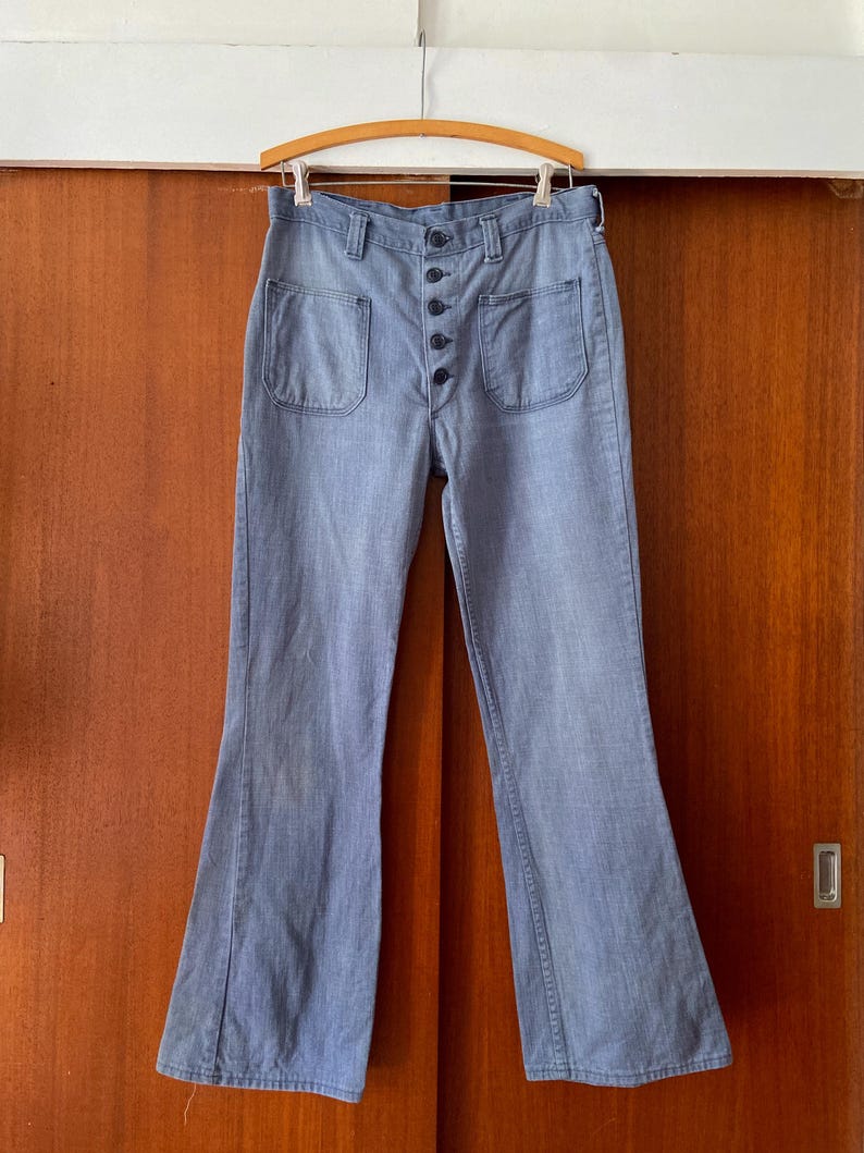 Puede incluir: Pantalones acampanados de mezclilla azul claro con bragueta abotonada y dos bolsillos delanteros. Los pantalones cuelgan de una percha de madera. La tela tiene un aspecto descolorido, lo que le da un aspecto vintage. Un estilo de pantal&oacute;n cl&aacute;sico.