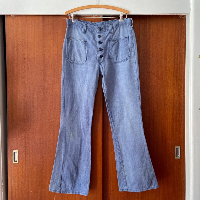 Puede incluir: Pantalones acampanados de mezclilla azul claro con grandes bolsillos delanteros y bragueta de botones. Los pantalones est&aacute;n colgados de una percha de madera. La mezclilla tiene un aspecto descolorido. Los pantalones est&aacute;n colgados frente a una puerta de madera.