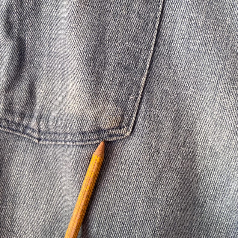 Puede incluir: Primer plano de una tela vaquera azul claro con un bolsillo y un l&aacute;piz amarillo y marr&oacute;n que apunta al bolsillo. El denim tiene un aspecto descolorido con algo de decoloraci&oacute;n.