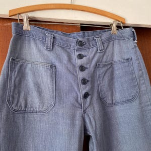 Puede incluir: Pantalones vaqueros azul claro con bolsillos delanteros grandes y cierre de botones. Los pantalones est&aacute;n colgados de una percha de madera. El denim tiene un aspecto descolorido.
