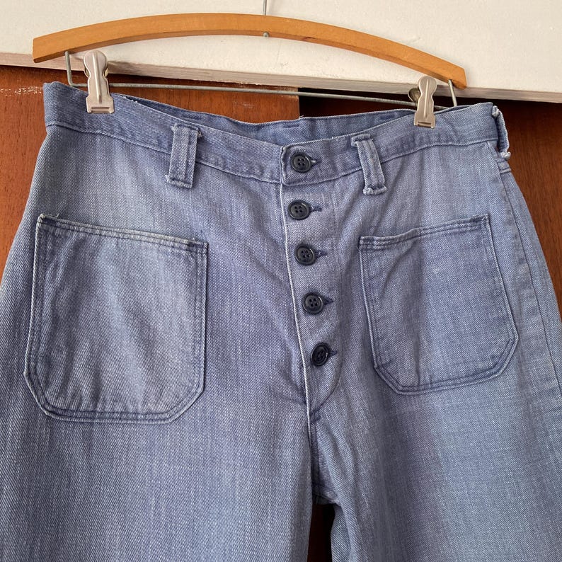 Puede incluir: Pantalones vaqueros azul claro con bolsillos delanteros grandes y cierre de botones. Los pantalones est&aacute;n colgados de una percha de madera. El denim tiene un aspecto descolorido.