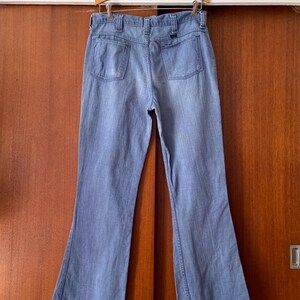 Puede incluir: Vaqueros de mezclilla azul claro con dise&ntilde;o de pierna acampanada, colgados de una percha de madera. Los vaqueros tienen dos bolsillos traseros y un estilo cl&aacute;sico de cinco bolsillos. La mezclilla parece ser de peso medio, adecuada para varias estaciones.