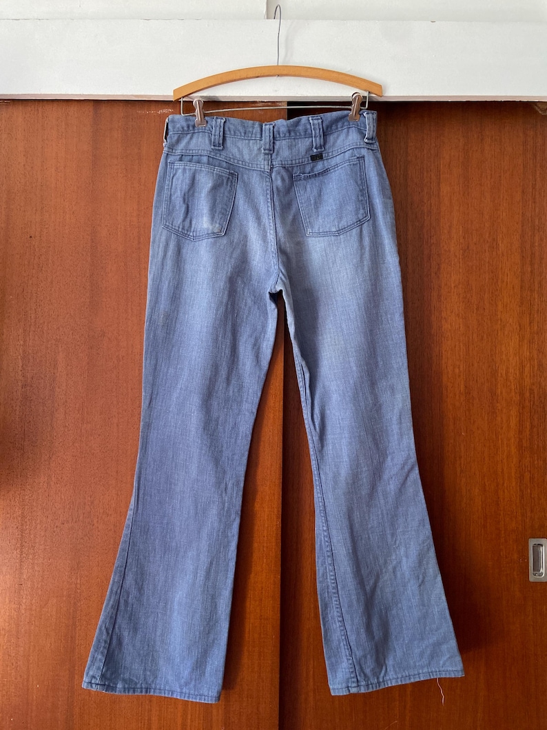 Puede incluir: Vaqueros de mezclilla azul claro con dise&ntilde;o de pierna acampanada, colgados de una percha de madera. Los vaqueros tienen dos bolsillos traseros y un estilo cl&aacute;sico de cinco bolsillos. La mezclilla parece ser de peso medio, adecuada para varias estaciones.