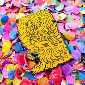 May include: A yellow enamel pin with a detailed illustration of a mythical creature with a snake tail and a single eye. The pin is surrounded by colorful confetti.