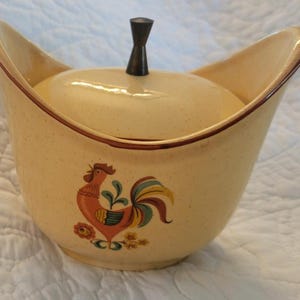 May include: A cream-colored ceramic sugar bowl with a speckled finish and a matching lid. The bowl features a brown rim and a central graphic of a rooster in orange, red, green, and blue. A dark brown, hourglass-shaped knob sits on the lid.