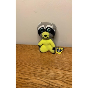 May include: A plush raccoon toy featuring a grey and white head with black eye patches, green eyes, and a bright yellow body. The toy has black whiskers and a small black nose. A blue and yellow tag with the text dream pets is attached to the side.