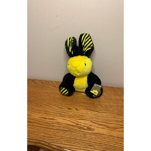 May include: A plush rabbit toy featuring a bright yellow face and chest contrasted with black fur on the limbs and back. The long ears display a black and yellow striped pattern. A small yellow tag with black text is attached to the right paw.