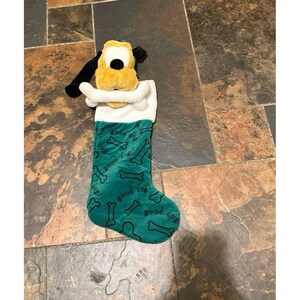 May include: A green Christmas stocking featuring a plush yellow dog head with black ears at the top, holding a white bone in its mouth. The stocking has a white faux fur cuff and is patterned with black outlines of bones and the text good dog.