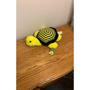 May include: A plush turtle toy featuring a bright yellow head and limbs with a black and yellow striped shell. The shell has a hexagonal pattern. A small rectangular tag with black and yellow diagonal stripes is attached to the front limb.