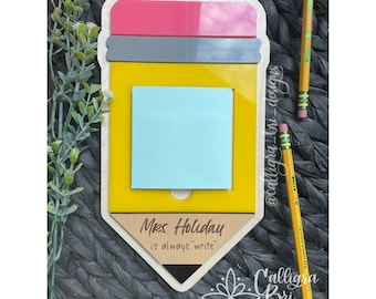 Pode incluir: Um sinal de madeira em forma de lápis com corpo amarelo, borracha rosa e grafite cinza. O sinal tem um recorte para um bloco de notas e o texto "Mrs. Holiday is always write" na parte inferior.