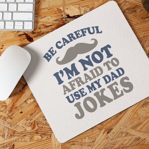 May include: A white mouse pad with the text "Be careful I'm not afraid to use my dad jokes" in blue and gray text. A gray mustache is above the text.