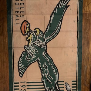 May include: A vintage-style banner with the words "EAGLES FOOTBALL" and the year "1933". The banner features a green and white eagle graphic holding a football. The background is tan with a green border.