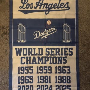Może przedstawiać: Niebiesko-biały baner Los Angeles Dodgers z nazwą drużyny, logo i latami mistrzostw World Series: 1955, 1959, 1963, 1965, 1981, 1988, 2020, 2024 i 2025. Baner ma oczka do zawieszenia.