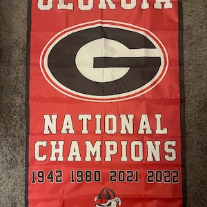 May include: Red banner with the word "GEORGIA" in white block letters at the top. Below is a black and white "G" logo inside a red and white oval. The words "NATIONAL CHAMPIONS" and the years 1942, 1980, 2021, and 2022 are printed below.