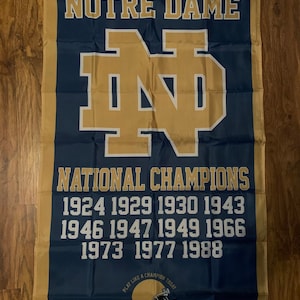 May include: A blue and gold Notre Dame flag with the school's logo and the text "NOTRE DAME" and "NATIONAL CHAMPIONS". The years of the school's national championships are listed below the text: 1924, 1929, 1930, 1943, 1946, 1947, 1949, 1966, 1973, 1977, 1988. The bottom of the flag has the text "PLAY LIKE A CHAMPION TODAY" and a gold football helmet.
