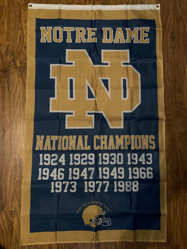Puede incluir: Una bandera azul y dorada de Notre Dame con el logotipo de la escuela y el texto "NOTRE DAME" y "NATIONAL CHAMPIONS". Los a&ntilde;os de los campeonatos nacionales de la escuela se enumeran debajo del texto: 1924, 1929, 1930, 1943, 1946, 1947, 1949, 1966, 1973, 1977, 1988. La parte inferior de la bandera tiene el texto "PLAY LIKE A CHAMPION TODAY" y un casco de f&uacute;tbol dorado.