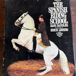 May include: A book cover titled "The Spanish Riding School" showing a white horse in a rearing pose with a rider in formal attire. The cover is black with text and an image of a horse and rider.