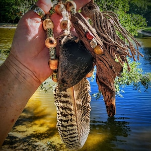 May include: A handcrafted, natural-themed pendant featuring a large, patterned feather, wooden beads, a dark, textured element, and a piece of driftwood. The pendant is adorned with small stones and twine, creating a rustic, earthy aesthetic.