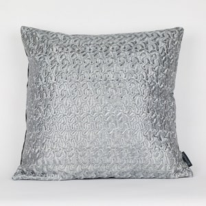 Può includere: Un cuscino da divano grigio argento con un motivo trapuntato testurizzato. Il cuscino ha un bordo nero e una piccola etichetta nera nell'angolo in basso a destra.