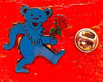 May include: A blue enamel pin shaped like a dancing bear holding a red rose with green leaves. The bear has black outlines and a smiling expression. Beside the pin is a circular metal clutch back fastener used to secure the pin to fabric.