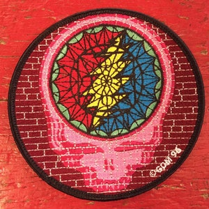 May include: A round, embroidered patch featuring a pink skull with a colourful, stained glass-like design inside. The skull is surrounded by a brick pattern and the text "GDW196" is visible in the lower right corner.