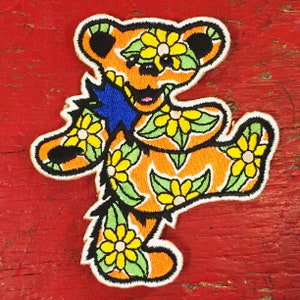 May include: Orange embroidered patch of a dancing bear with a blue ribbon around its neck. The bear is covered in yellow flowers and green leaves.