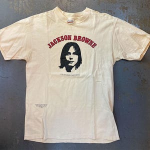 May include: Cream-coloured t-shirt with the text "JACKSON BROWNE" in red arched lettering above a black and white portrait. Below the portrait, the text reads "LOS ANGELES CALIFORNIA". The shirt has short sleeves and a crew neck.