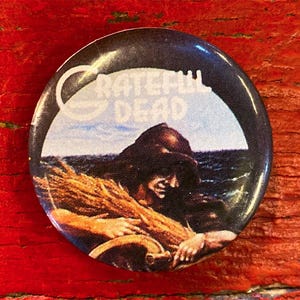 May include: A round button featuring a black and white image of a person holding a bundle of wheat. The text "GRATEFUL DEAD" is printed in white on a blue sky background.