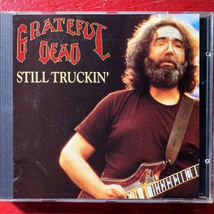 May include: CD case for the Grateful Dead's "Still Truckin'" album. The cover features a man with long hair and a beard playing a guitar. The band's name is in red gothic lettering above the album title.