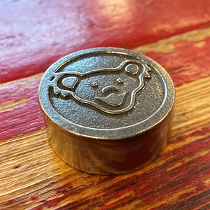 Puede incluir: Un recipiente de metal plateado con una tapa redonda que presenta un diseño de cara de oso. El recipiente está sentado sobre una superficie de madera con pintura roja.