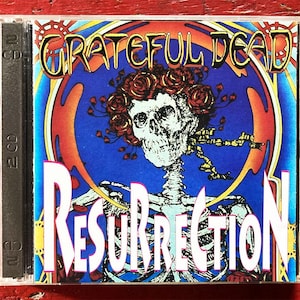 Può includere: Custodia di un CD dell'album "Resurrection" dei Grateful Dead. La copertina presenta uno scheletro con una corona di rose su sfondo blu. Il nome della band è dorato e il titolo dell'album è bianco.