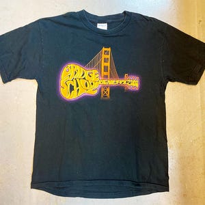 Könnte beinhalten: Schwarzes T-Shirt mit einem Aufdruck einer Gitarre und der Golden Gate Bridge. Der Text "Bridge School Benefit 2004" ist auf dem Shirt gedruckt.
