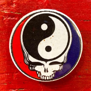 May include: A round button with a yin yang symbol in black and white, with a red and blue background. The yin yang symbol is surrounded by a cartoon skull with a red and blue background.