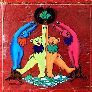 May include: A colorful sticker featuring dancing bears in blue, yellow, orange, and pink, with peace signs and a maple leaf. The bears are standing on a patch of grass and rocks, with a red background.